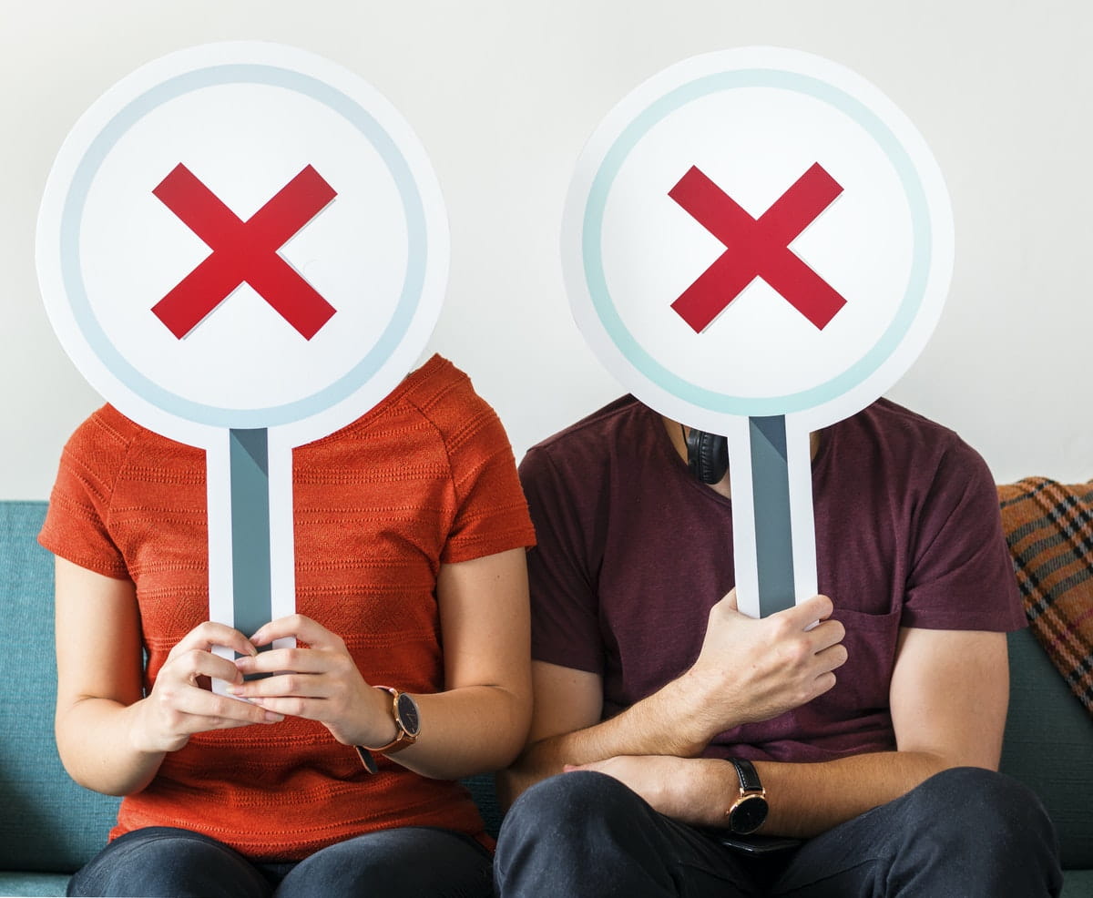 Two people sitting, holding white circles with red X's over their faces